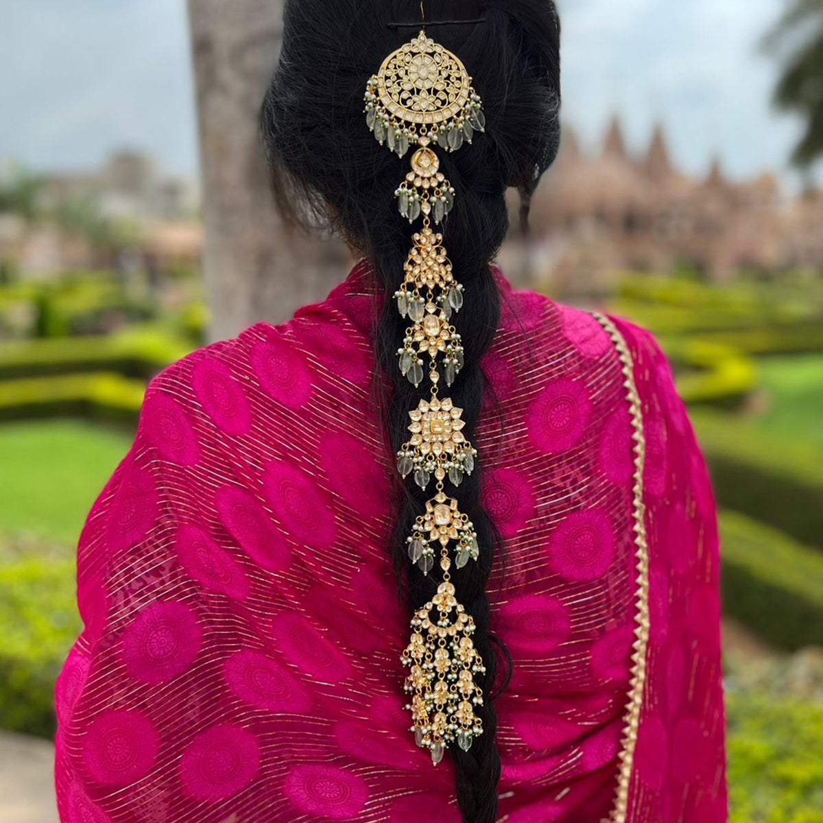 A gold-plated kundan bridal hair accessory adorning a braid, with semi-precious stones and natural gemstones, worn by a person in a pink saree.