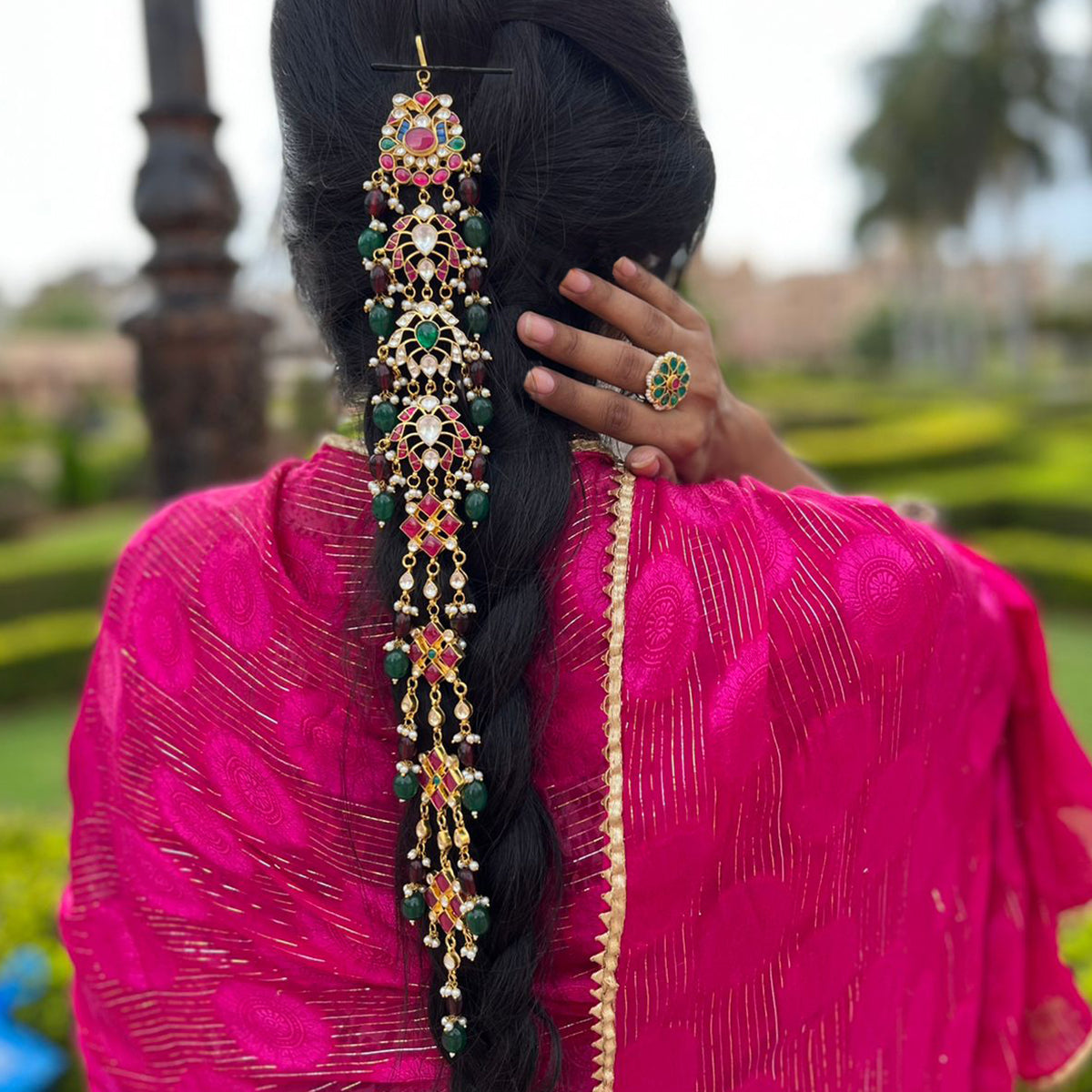 A person wearing a traditional pink saree with a gold-plated and multicolored kundan hair accessory in their braided hair.
